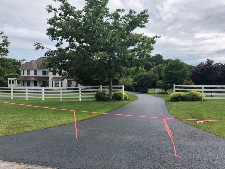 Paved driveway blocked by bright orange caution tape across the path, with a white fence, green lawn, and a two-story house in the background.