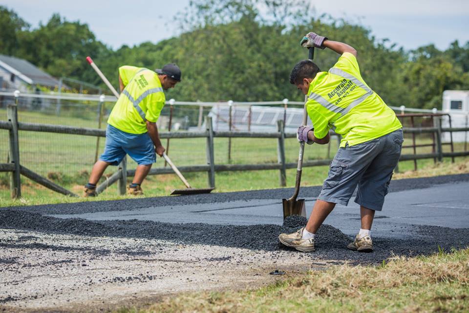 Two construction workers in neon vests spread and smooth fresh asphalt on a rural road.